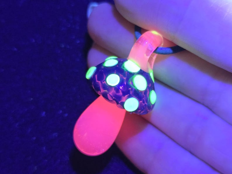 UV-reactive borosilicate glass mushroom pendant glowing fluorescent green and pink under blacklight, showing dramatic color transformation from its natural blue tones.