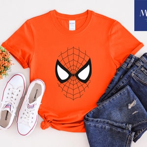Pu&ograve; includere: Una maglietta arancione con un disegno a maschera di Spider-Man in bianco e nero. L'immagine mostra la maglietta insieme a jeans blu e scarpe da ginnastica bianche. Sono visibili anche una piccola composizione floreale e un logo di un negozio di stampa.