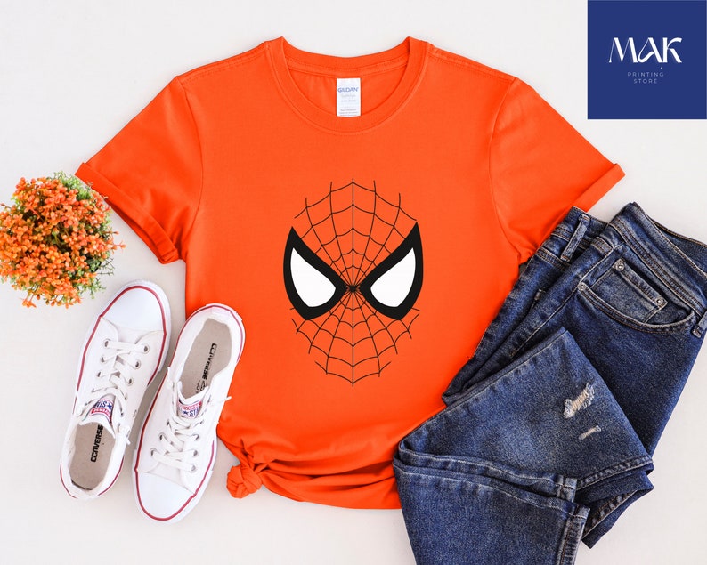 Pu&ograve; includere: Una maglietta arancione con un disegno a maschera di Spider-Man in bianco e nero. L'immagine mostra la maglietta insieme a jeans blu e scarpe da ginnastica bianche. Sono visibili anche una piccola composizione floreale e un logo di un negozio di stampa.