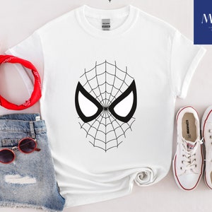 Pu&ograve; includere: T-shirt bianca con un design a maschera di Spider-Man in bianco e nero. L'outfit include pantaloncini di jeans, occhiali da sole rossi, una bandana rossa e scarpe da ginnastica bianche. Il logo "MAK PRINTING STORE" &egrave; visibile.