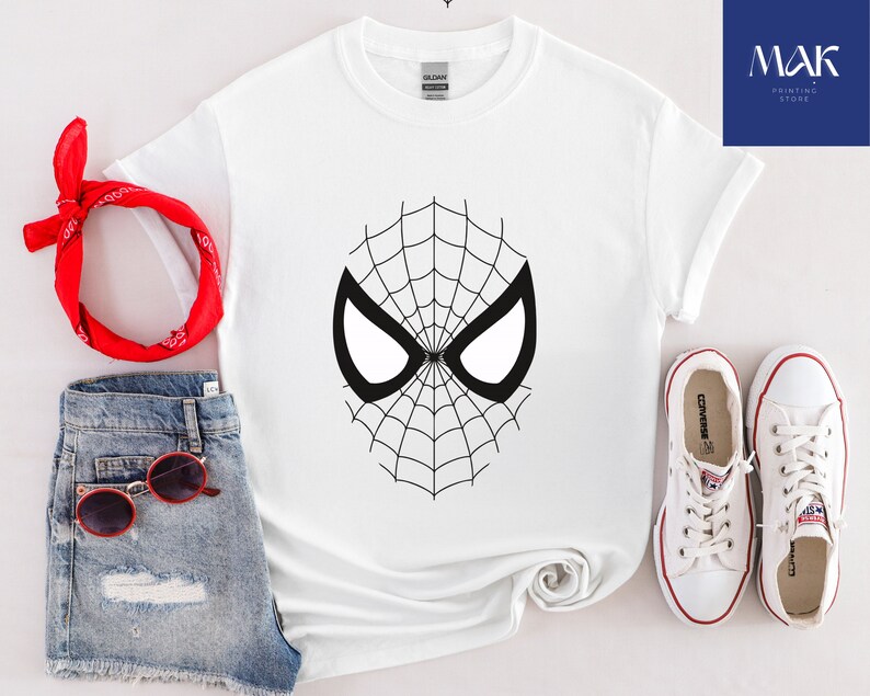 Pu&ograve; includere: T-shirt bianca con un design a maschera di Spider-Man in bianco e nero. L'outfit include pantaloncini di jeans, occhiali da sole rossi, una bandana rossa e scarpe da ginnastica bianche. Il logo "MAK PRINTING STORE" &egrave; visibile.