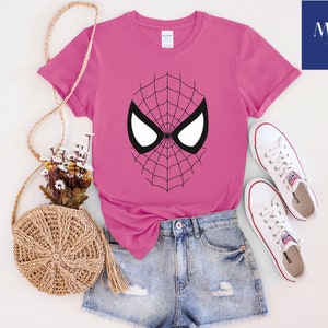 Pu&ograve; includere: Una maglietta magenta con un design a maschera di Spider-Man in bianco e nero. La maglietta &egrave; abbinata a pantaloncini di jeans e scarpe da ginnastica bianche. &Egrave; visibile anche una borsa intrecciata con fiori e la scritta "MAK PRINTING STORE".