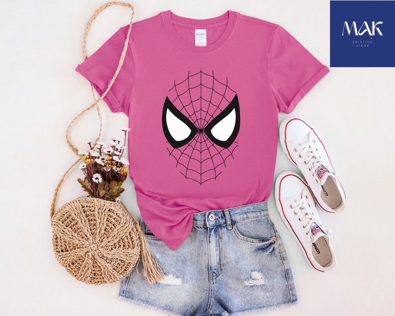 Pu&ograve; includere: Una maglietta magenta con un design a maschera di Spider-Man in bianco e nero. La maglietta &egrave; abbinata a pantaloncini di jeans e scarpe da ginnastica bianche. &Egrave; visibile anche una borsa intrecciata con fiori e la scritta "MAK PRINTING STORE".