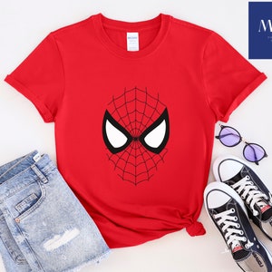 Pu&ograve; includere: Una maglietta rossa con un disegno a maschera di Spider-Man in bianco e nero. L'immagine include pantaloncini di jeans blu, scarpe da ginnastica nere e occhiali da sole rotondi. &Egrave; visibile anche il testo "MAK Printing Store".