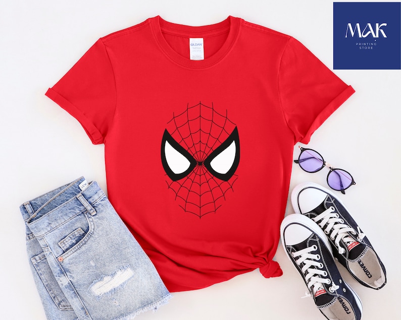 Pu&ograve; includere: Una maglietta rossa con un disegno a maschera di Spider-Man in bianco e nero. L'immagine include pantaloncini di jeans blu, scarpe da ginnastica nere e occhiali da sole rotondi. &Egrave; visibile anche il testo "MAK Printing Store".