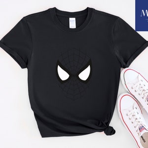 Pu&ograve; includere: T-shirt nero con grafica della maschera di Spider-Man. Il design include forme di occhi bianchi e un motivo a ragnatela nero. Scarpe da ginnastica bianche con dettagli rossi sono accanto alla maglietta. La maglietta &egrave; di MAK Printing Store.