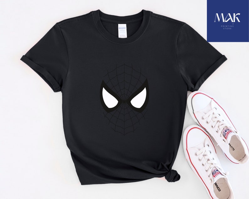Pu&ograve; includere: T-shirt nero con grafica della maschera di Spider-Man. Il design include forme di occhi bianchi e un motivo a ragnatela nero. Scarpe da ginnastica bianche con dettagli rossi sono accanto alla maglietta. La maglietta &egrave; di MAK Printing Store.