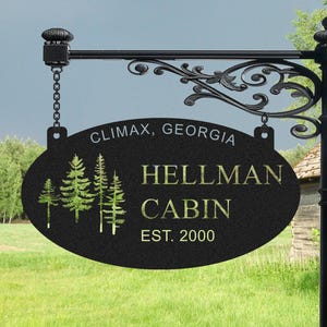 May include: Black oval sign with gold text that reads "Hellman Cabin Est. 2000" and "Climax, Georgia". The sign has a silhouette of pine trees and is hanging from a black metal bracket.