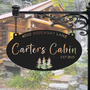 May include: Black oval sign with white text that reads "4000 Hideaway Lane Carters Cabin Est 2025". The sign is hanging from a black metal bracket with a chain. The sign has a silhouette of three pine trees in the centre.