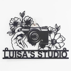 Pode incluir: Um letreiro de metal preto com uma câmera e um design floral. O letreiro diz "LUISA'S STUDIO".