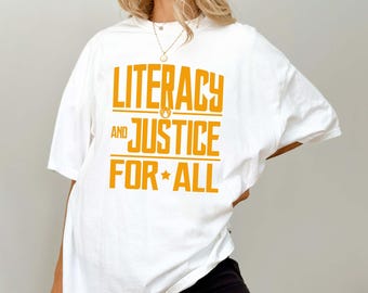 May include: White t-shirt with the text "LITERACY AND JUSTICE FOR ALL" in orange block letters. The shirt has short sleeves and a relaxed fit. The text includes a star graphic and a flame graphic.