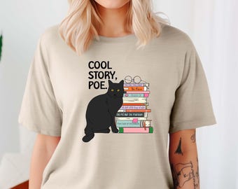 May include: Beige t-shirt featuring a black cat illustration sitting next to a stack of books. The text "COOL STORY, POE." is printed above the cat. The books have titles like "The Raven" and "The Pit and the Pendulum".