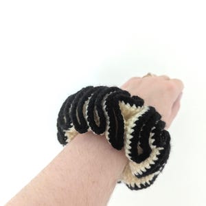 Pu&ograve; includere: Un elastico per capelli all'uncinetto nero e crema. L'accessorio per capelli &egrave; realizzato in filato e presenta un motivo ondulato e strutturato. L'elastico &egrave; indossato al polso.