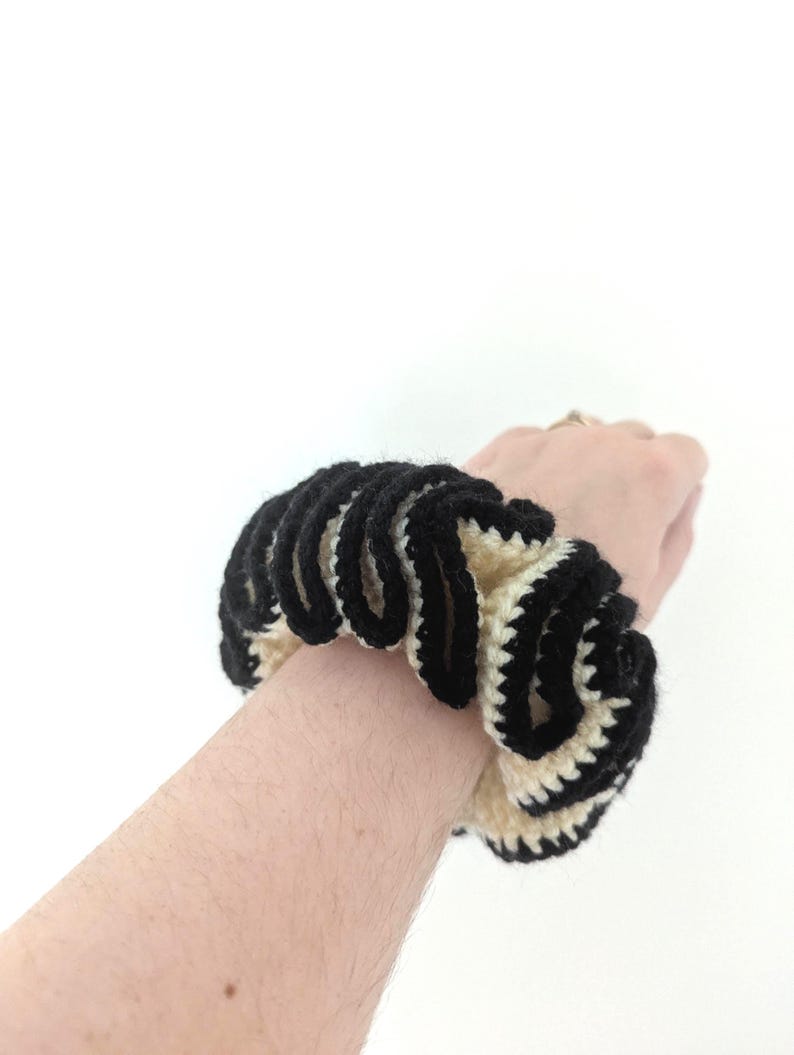 Pu&ograve; includere: Un elastico per capelli all'uncinetto nero e crema. L'accessorio per capelli &egrave; realizzato in filato e presenta un motivo ondulato e strutturato. L'elastico &egrave; indossato al polso.