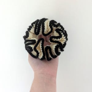Pu&ograve; includere: Un elastico per capelli fatto a mano con una base color crema e un bordo nero ondulato. L'elastico &egrave; realizzato in un materiale morbido e strutturato ed &egrave; progettato per tenere i capelli. L'elastico &egrave; tenuto in mano su sfondo bianco.