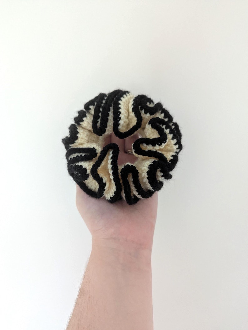 Pu&ograve; includere: Un elastico per capelli fatto a mano con una base color crema e un bordo nero ondulato. L'elastico &egrave; realizzato in un materiale morbido e strutturato ed &egrave; progettato per tenere i capelli. L'elastico &egrave; tenuto in mano su sfondo bianco.