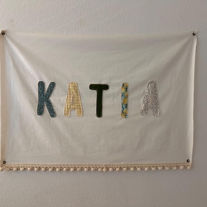 Peut inclure: Une bannière en tissu blanc avec le nom "KATIA" écrit en lettres de tissu colorées. La bannière est ornée d'une bordure de pompons blancs.