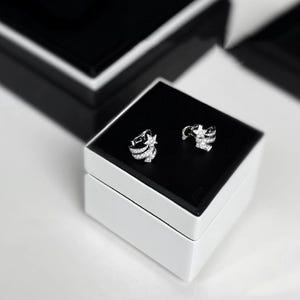 May include: A pair of silver star-shaped earrings with clear stones, displayed in a white and black box.