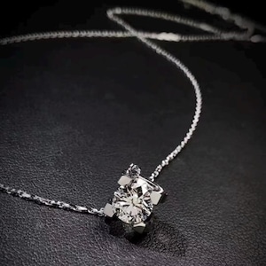 Könnte beinhalten: Eine zarte Silberkette mit einem funkelnden, rund geschliffenen Diamantanhänger. Der Diamant wird von einer Vier-Krappen-Fassung gehalten, und die Kette ist fein und elegant. Die Halskette wird vor einem dunklen Hintergrund präsentiert.