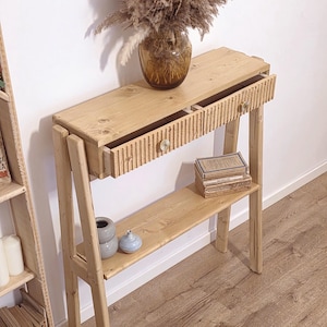 May include: A light-coloured wooden console table with a drawer and a lower shelf. The drawer has a ribbed design and a decorative knob. A vase with dried flowers sits on top. The table is 102 cm long.