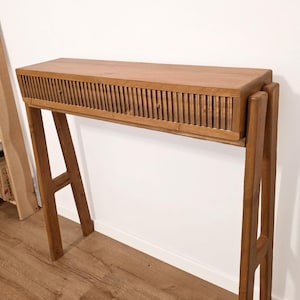Op de afbeelding: Een houten console in mid-century modern stijl. De tafel heeft een rechthoekig blad met een lattenfront en schuine poten. Het hout heeft een warme, natuurlijke afwerking. De tafel is ongeveer 122 cm lang.