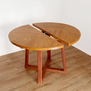 Könnte beinhalten: Ein zweiteiliger, halbkreisförmiger Holztisch mit warmbraunem Finish. Der Tisch steht auf einem passenden Holzfuß mit einem kreuzbeinigen Design. Der Tisch steht auf einem hellbraunen Holzboden.