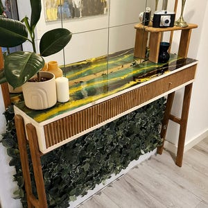 Può includere: Una consolle in legno con piano in resina dai toni gialli e verdi. Il tavolo ha un frontale del cassetto a coste e gambe in legno angolate. Una pianta e candele sono sul tavolo. Uno specchio è sullo sfondo.