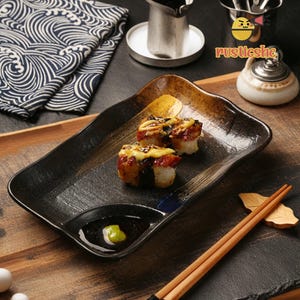 Peut inclure: Une assiette rectangulaire en céramique noire et dorée avec deux morceaux de sushi dessus. Les sushis sont recouverts d'une sauce jaune et de graines de sésame noires. Il y a un petit plat avec de la sauce verte sur l'assiette. Une paire de baguettes en bois est au premier plan.