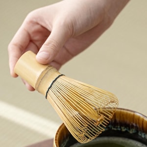 May include: A bamboo matcha whisk with a natural wood handle. The whisk is being held over a bowl of green matcha tea.