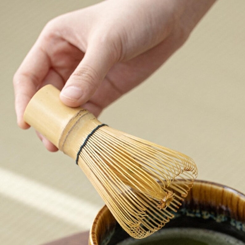 May include: A bamboo matcha whisk with a natural wood handle. The whisk is being held over a bowl of green matcha tea.