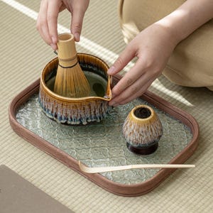 May include: A brown and white ceramic tea bowl with a bamboo whisk and a small brown and white ceramic cup on a tray with a light blue and white patterned surface.