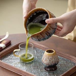 May include: A brown and white ceramic tea pitcher pouring green tea into a clear glass mug. The tea pitcher and mug are on a wooden tray with a decorative pattern.