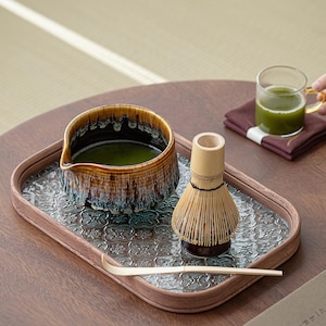May include: A wooden tray with a patterned surface holds a ceramic bowl filled with green matcha tea, a bamboo whisk, and a wooden spoon. The tray is on a wooden table.