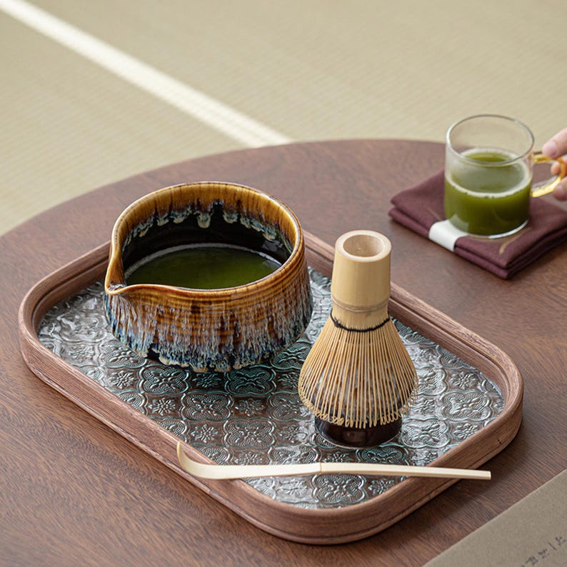 May include: A wooden tray with a patterned surface holds a ceramic bowl filled with green matcha tea, a bamboo whisk, and a wooden spoon. The tray is on a wooden table.