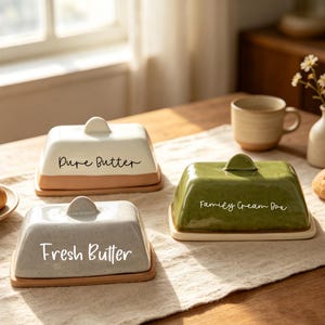 May include: Three rectangular ceramic butter dishes with lids. One is cream with 'Pure Butter' text, one is light grey with 'Fresh Butter' text, and one is olive green with 'Family Cream Box' text. Each dish has a matching base and a small handle.