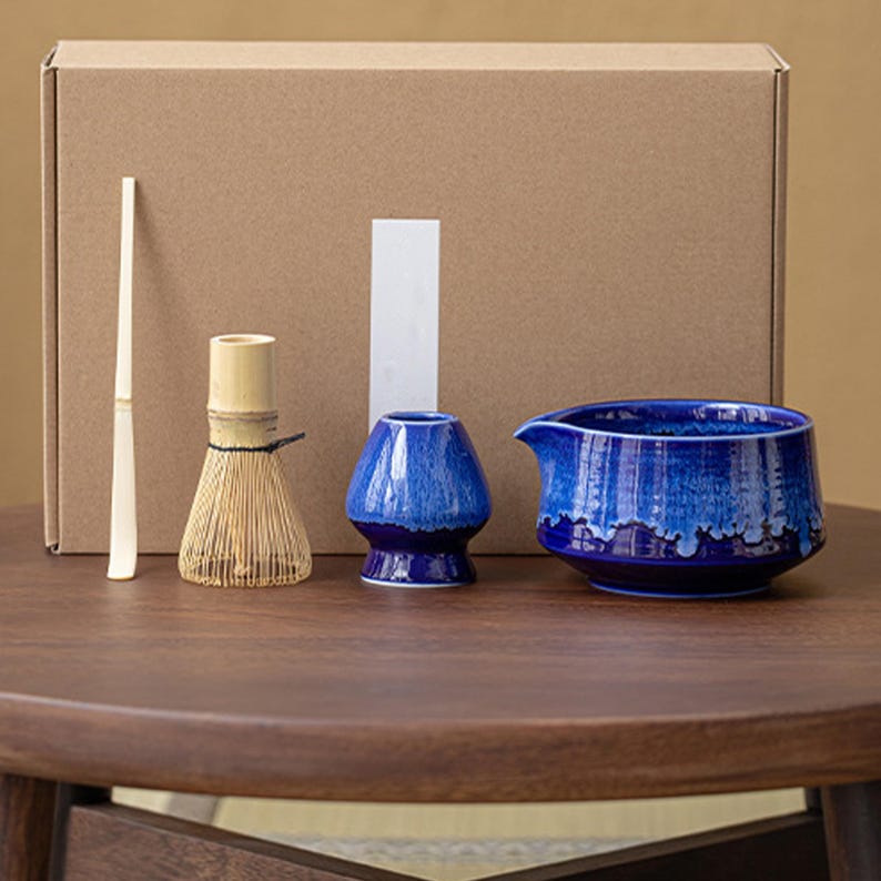 May include: A blue and white ceramic matcha set with a whisk, a bowl, and a pitcher. The set is arranged on a wooden table.