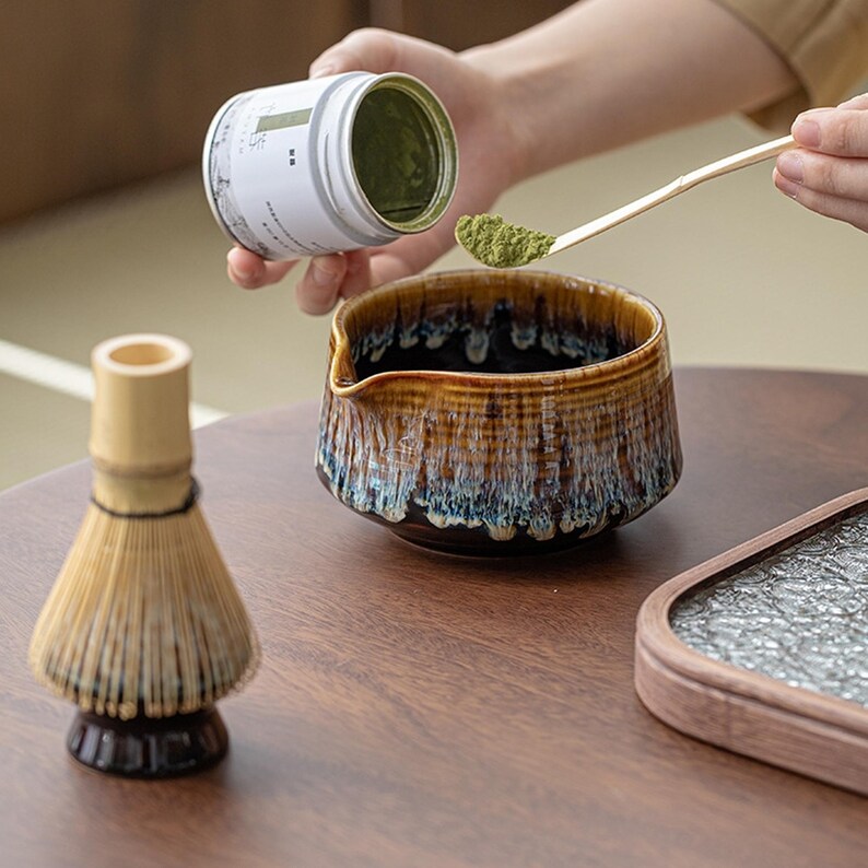 May include: A brown and white ceramic tea bowl with a blue and white design. A person is pouring green tea powder from a white container into the bowl. A bamboo whisk and a wooden tray are also visible.