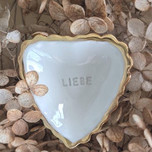 Könnte beinhalten: Eine weiße, herzförmige Schale mit goldenem Rand und dem Wort "LIEBE" auf der Oberfläche. Die Schale ist von getrockneten, hellbraunen Blüten umgeben, was einen weichen, natürlichen Hintergrund erzeugt.