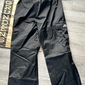 Puede incluir: Pantal&oacute;n impermeable negro con cintura el&aacute;stica naranja y negra y un logotipo blanco que dice "Grundens" en la pierna derecha.