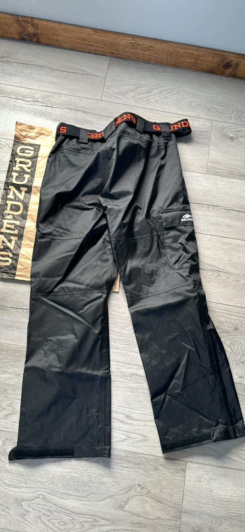 Puede incluir: Pantal&oacute;n impermeable negro con cintura el&aacute;stica naranja y negra y un logotipo blanco que dice "Grundens" en la pierna derecha.