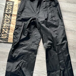 Puede incluir: Pantal&oacute;n cargo negro con cintur&oacute;n negro y texto naranja que dice "GRUNDENS" en la pierna izquierda y "NDE" en la pierna derecha.