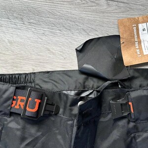 Puede incluir: Pantal&oacute;n de esqu&iacute; negro con una cintura el&aacute;stica naranja y negra. La cintura tiene las palabras "GR&Uuml;NDER" e "INS" impresas en ella.