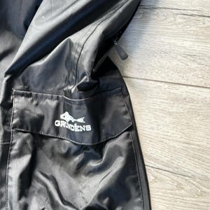 Puede incluir: Pantal&oacute;n de pesca negro con un bolsillo grande en el lado derecho. El bolsillo tiene un logotipo blanco que dice "Grundens" con un gr&aacute;fico de pescado. El pantal&oacute;n tiene una correa ajustable naranja y negra.