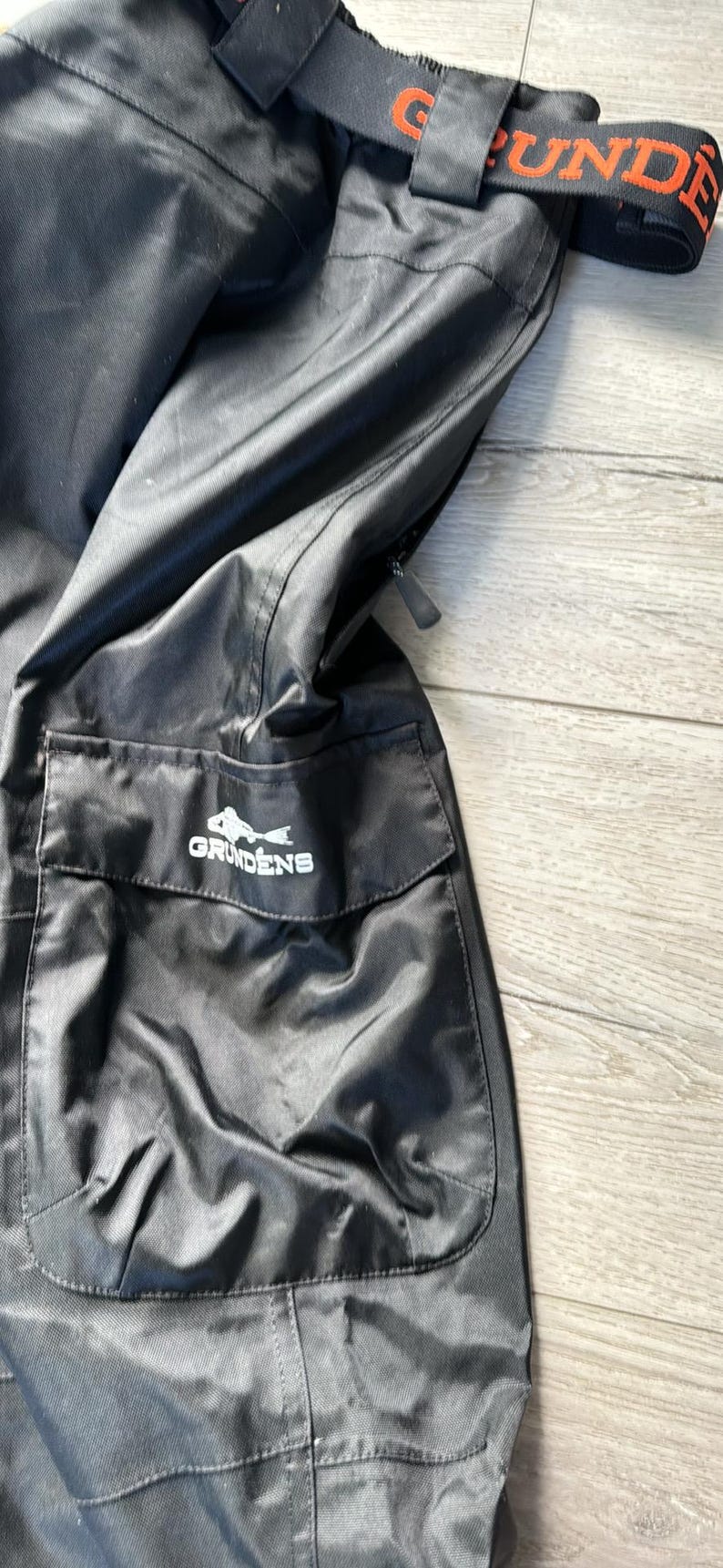 Puede incluir: Pantal&oacute;n de pesca negro con un bolsillo grande en el lado derecho. El bolsillo tiene un logotipo blanco que dice "Grundens" con un gr&aacute;fico de pescado. El pantal&oacute;n tiene una correa ajustable naranja y negra.