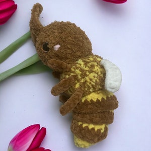 May include: A crocheted plush toy shaped like a bee with brown and yellow fur. The bee has a white wing and large brown eyes. The bee is lying on a white surface with pink tulips.