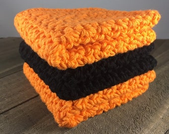 May include: A stack of three crocheted dishcloths in orange and black. The dishcloths are made with a textured stitch.