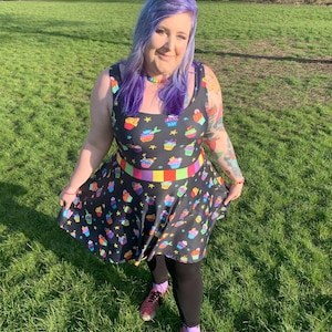 May include: A woman with purple hair is wearing a black skater dress with a cupcake print. The dress has a rainbow stripe around the waist. She is standing in a grassy field.