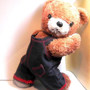 Op de afbeelding: Een bruine teddybeer die een zwarte denim overall draagt met rode stiksels en rode bies. De beer staat op zijn achterpoten met zijn voorpoten omhoog.