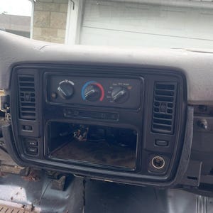 May include: A black automotive dashboard centre console panel featuring three circular climate control knobs, vertical air vents on both sides, a rectangular opening for a radio unit, and buttons labelled E/M and TRIP on the left side.