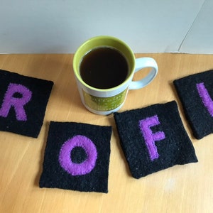May include: Four black square coasters with purple letters spelling "ROFL" are arranged on a wooden surface. A mug with a green and white design and filled with a dark liquid sits nearby. The coasters are made of a textured material.